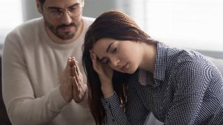 A woman with her head in her hand while a man sitting behind her has his hands together in a praying motion, asking for forgiveness.