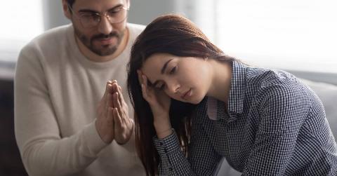 A woman with her head in her hand while a man sitting behind her has his hands together in a praying motion, asking for forgiveness.