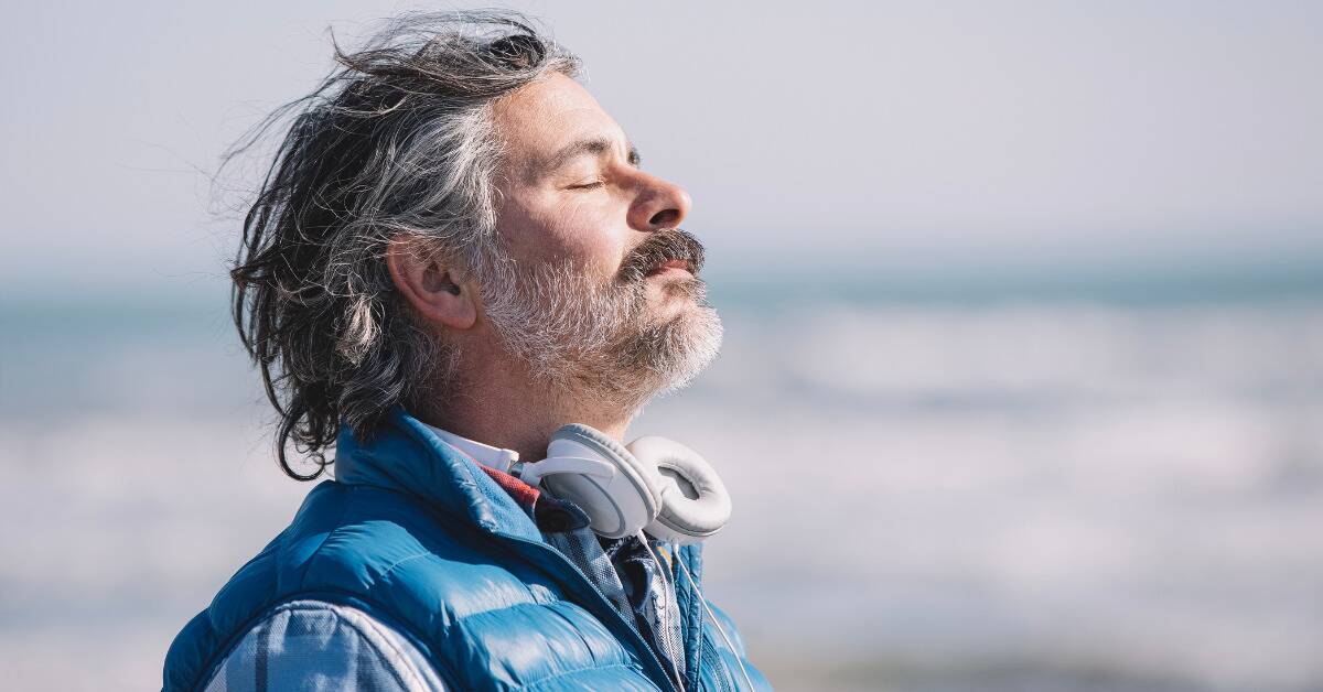 A man walking on the beach, eyes closed, head tilted up, the wind blowing his hair back.