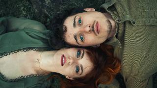 A couple laying outside, their heads side by side but their bodies in opposite directions, looking up at the camera with intense eyes.