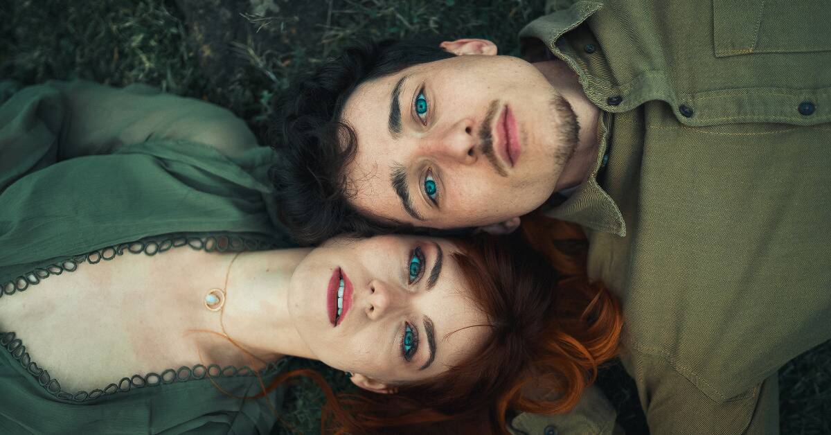 A couple laying outside, their heads side by side but their bodies in opposite directions, looking up at the camera with intense eyes.