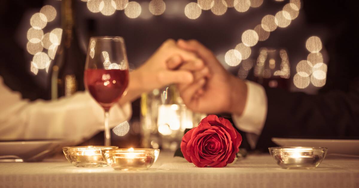 A couple holding hands across a table, a glass of wine and candles also visible, the camera focused on a rose placed in the middle of it all.