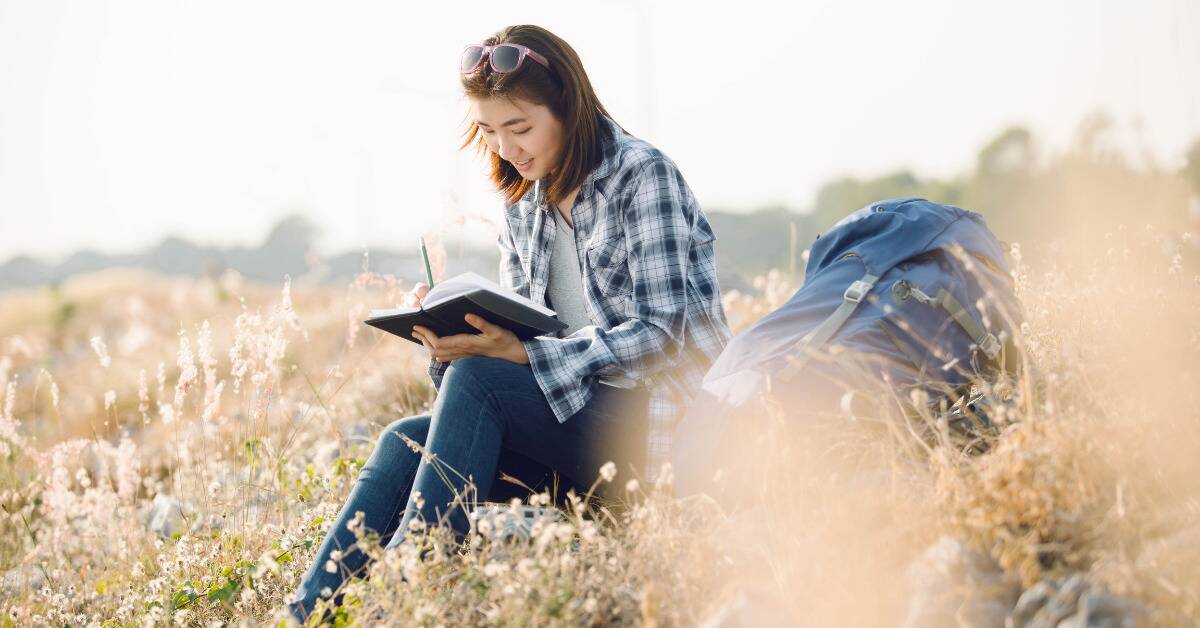 A woman sitting in some tall grass, smiling as she writes in her journal.