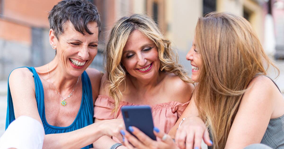 Three women all smiling outside as they all look at something on one of their phones.