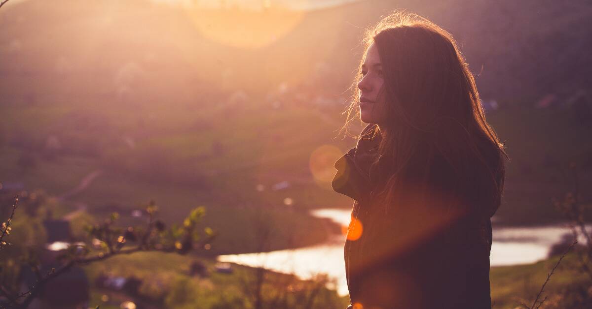 A woman standing outside during a sunrise, looking off into the distance contemplatively.