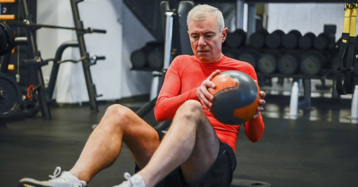 A man in the gym, dong oblique twists while holding a medicine ball.