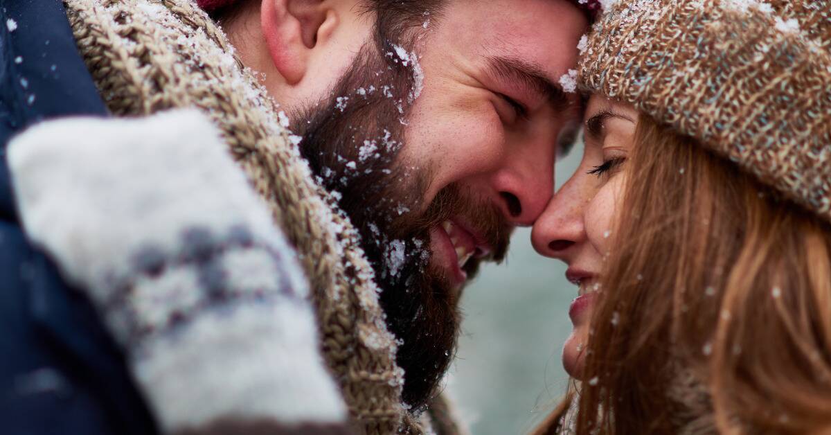A close shot of a couple with their noses pressed together outside in the winter, their eyes closed, both smiling.