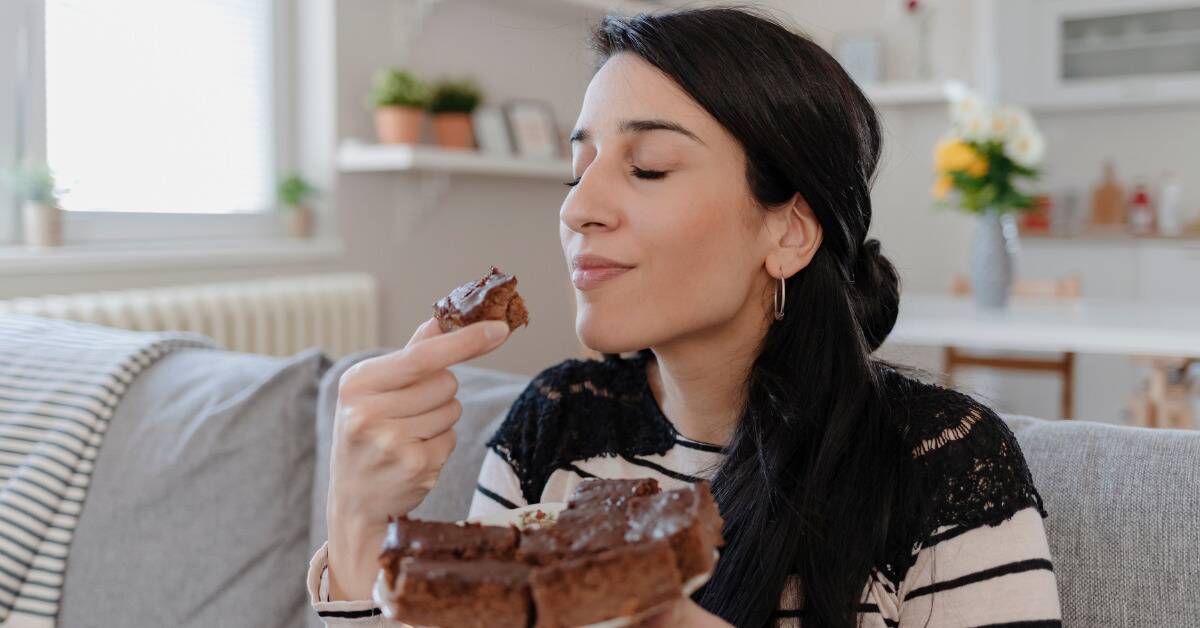 A woman holding a plate of brownies with one to her mouth, her eyes closed as she smiles, having just taken a bite.