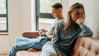A man sitting on the couch next to her boyfriend who's on his phone, she looks annoyed and exasperated, facing away from him.