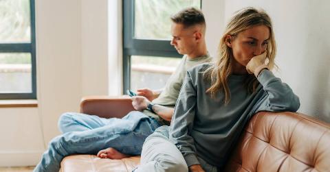 A man sitting on the couch next to her boyfriend who's on his phone, she looks annoyed and exasperated, facing away from him.