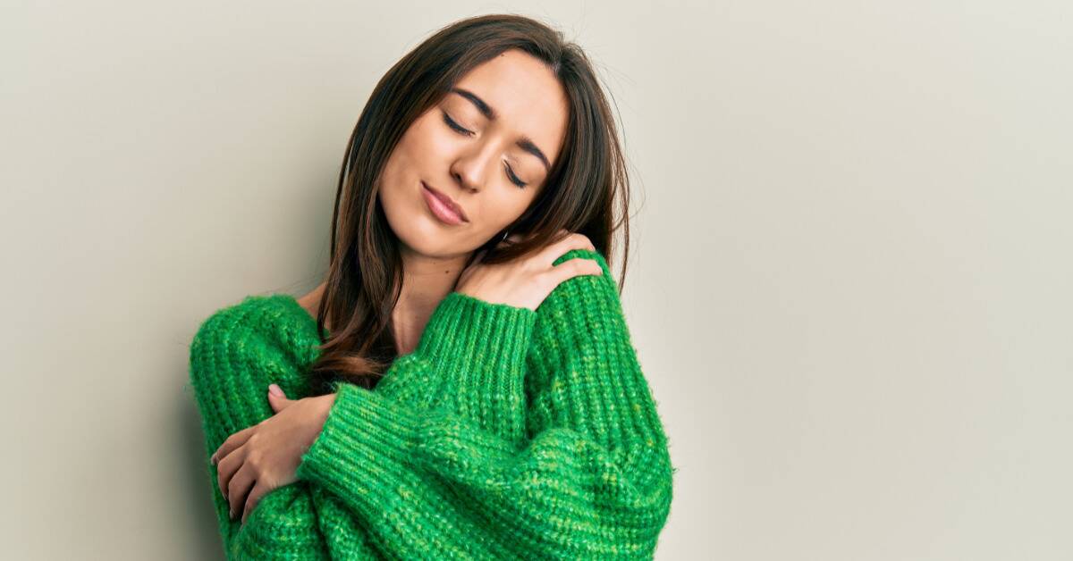 A woman in a bright green sweater smiling with her eyes closed as she hugs herself.