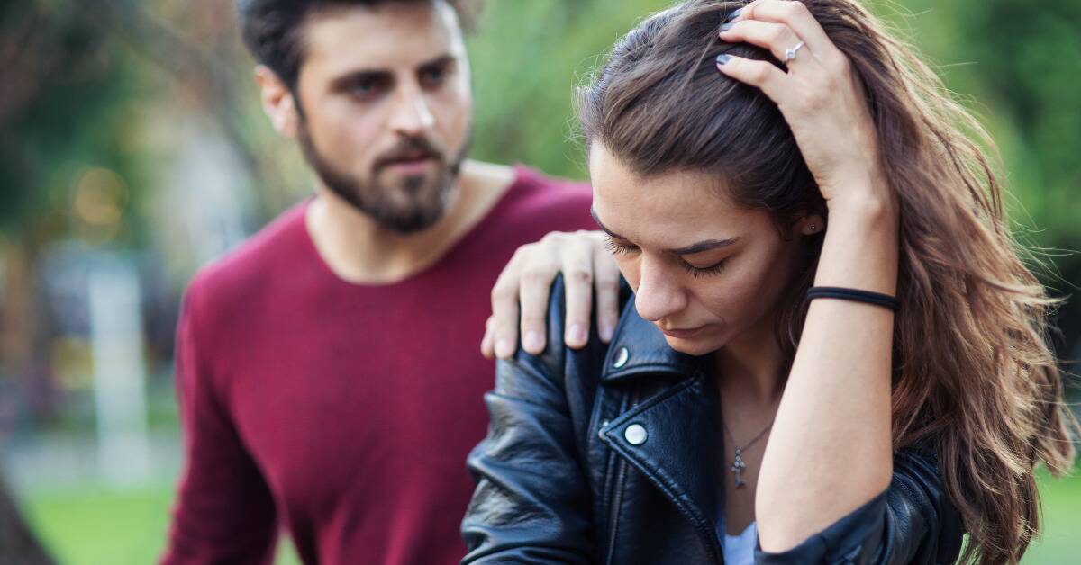 A woman looking sad, a man behind her seen trying to talk to her, his hand on her shoulder.