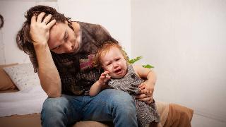 A dad holding his crying baby, head in his hand, looking stressed.