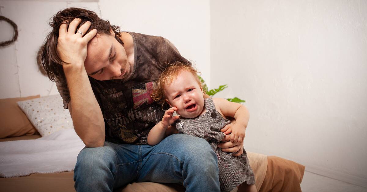 A dad holding his crying baby, head in his hand, looking stressed.