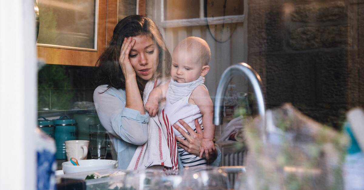 A mom holding her baby, hand on her face, looking stressed.