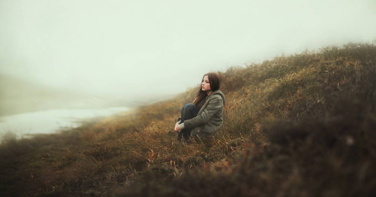 A woman sitting along on a grassy, foggy hill.