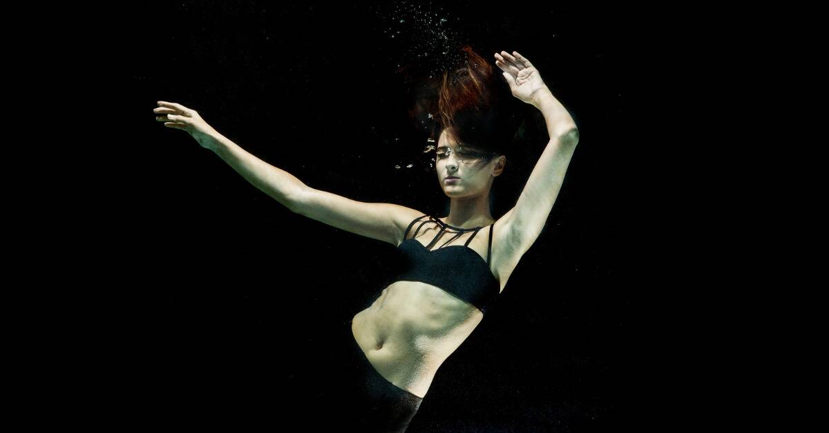 A woman seeming to be freefalling underwater, arms limp and raising upwards, bubbles coming from her nose.