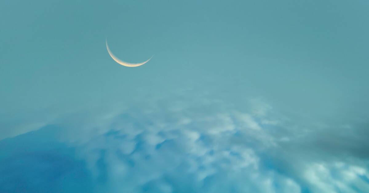 A thin crescent moon above a layer of light blue clouds.