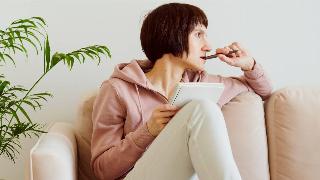 A woman sitting on a couch, one leg drawn up, resting a journal on her knee, looking off to the side and chewing on her pen as she thinks.