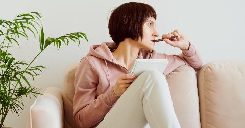 A woman sitting on a couch, one leg drawn up, resting a journal on her knee, looking off to the side and chewing on her pen as she thinks.
