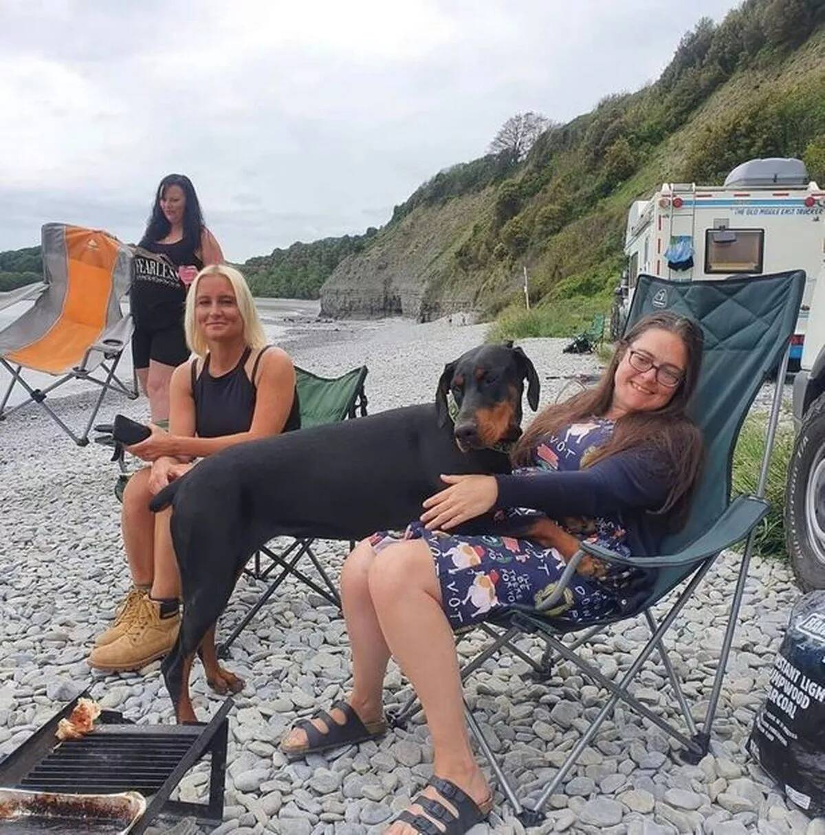 Lucy and Katie sitting in foldout chairs on the beach, Indie the doberman half in Lucy's lap.