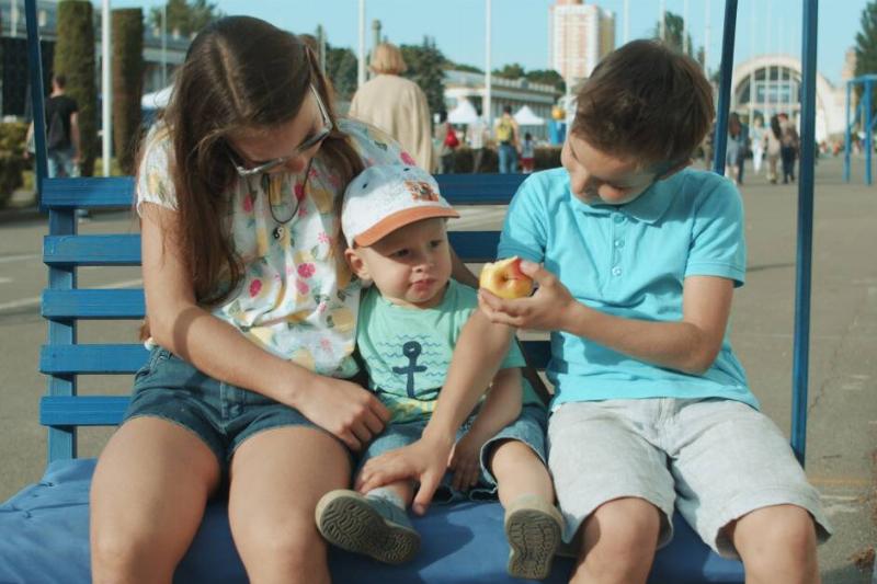 Two older siblings sitting with their baby brother on a bench, trying to feed him an apple.