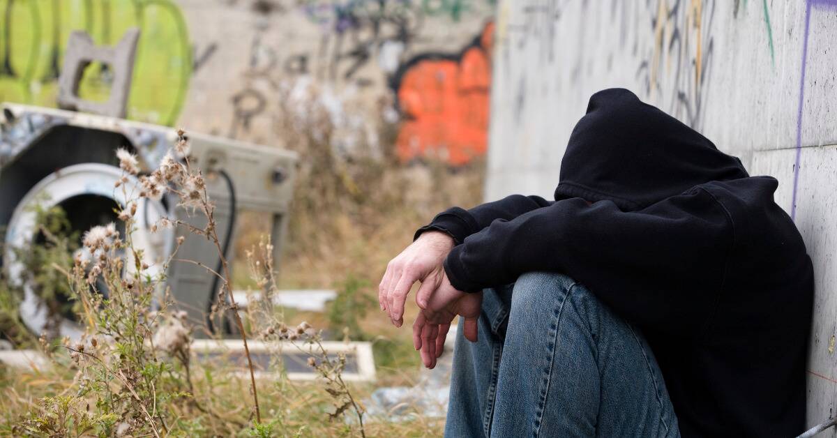 A teen sitting in an abandoned industrial area, hood pulled up and knees drawn to his chest, hiding his face.
