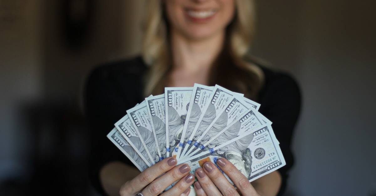 A woman smiling as she holds out a fan of $100 bills.