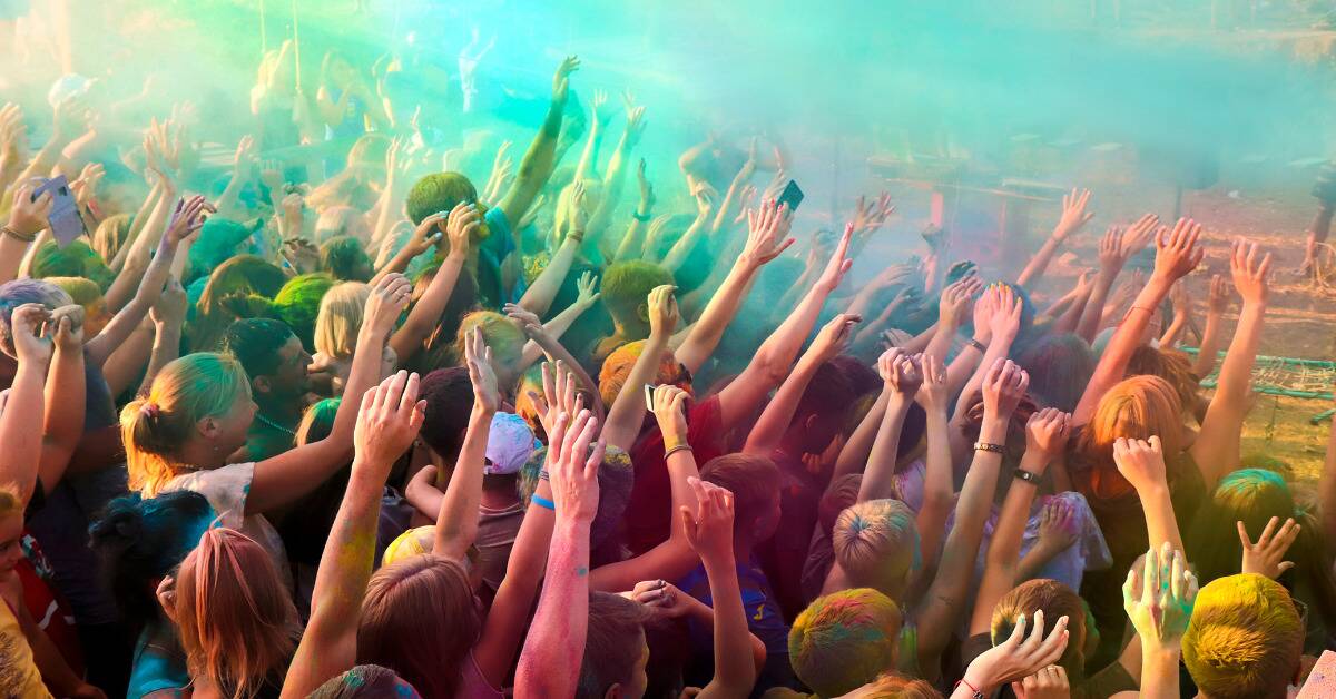 A crowd raising their hands as colorful sand is thrown around at a festival.