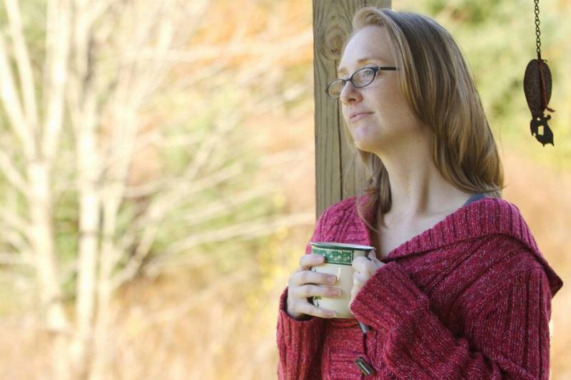 A woman standing outside with a mug of coffee, looking into the distance peacefully.