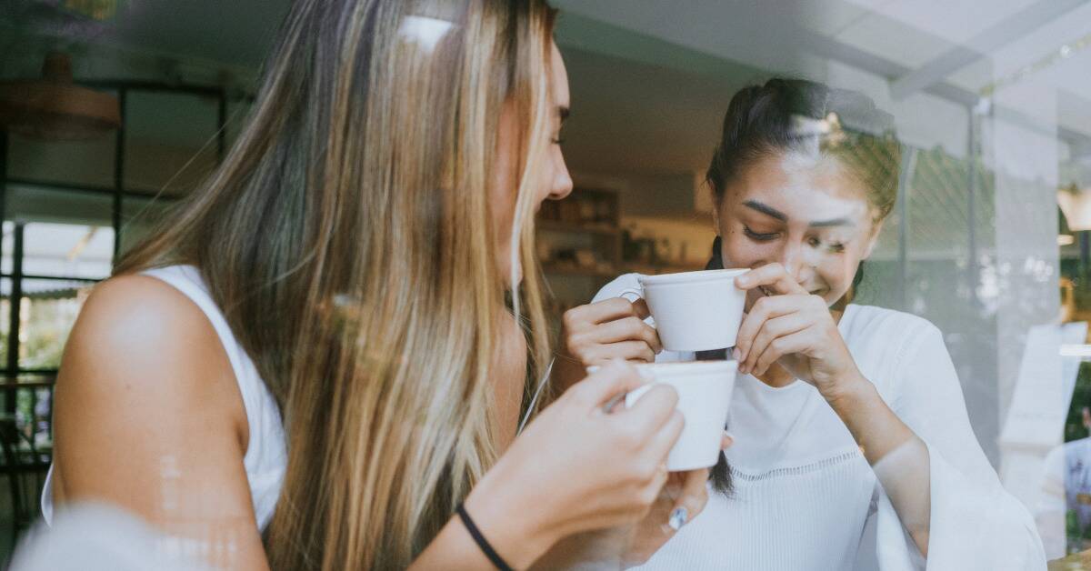 Two friends laughing and chatting over cups of coffee at a cafe.
