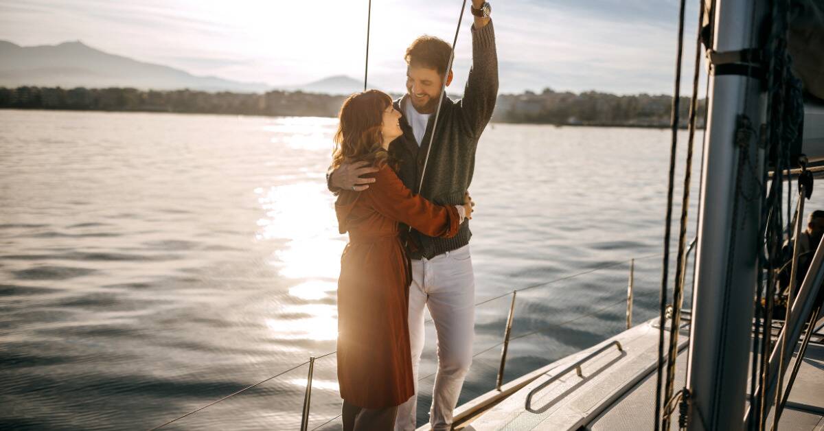 A couple hugging and smiling as they stand on a boat, the man holding onto a bar above his head.