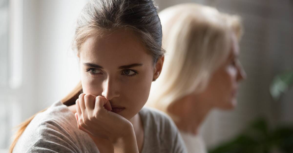 A woman with her chin in her hand, looking cold and angry, looking away from her mother who's visible blurred in the background.