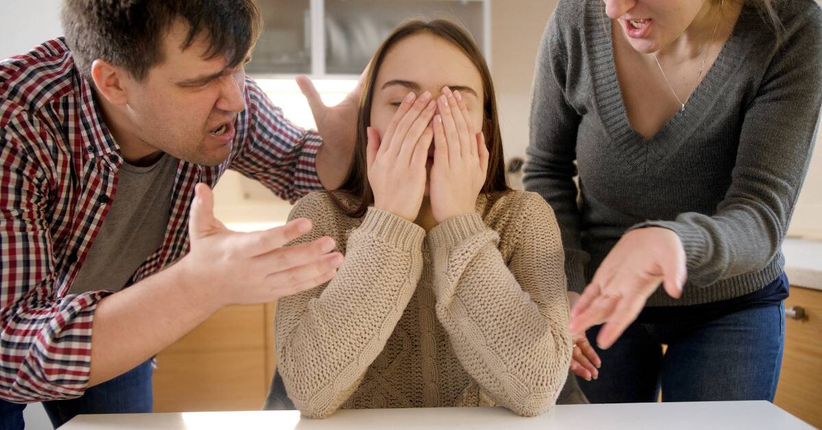 A woman sitting at a table, covering her face with her hands, both parents on either side of her yelling at her.