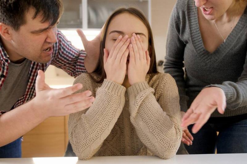 A woman sitting at a table, covering her face with her hands, both parents on either side of her yelling at her.