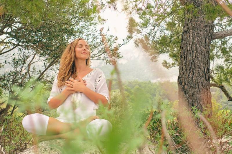 A woman sitting in a wooded area, legs crossed in a meditation pose, eyes closed, hands over her heart, smiling peacefully.