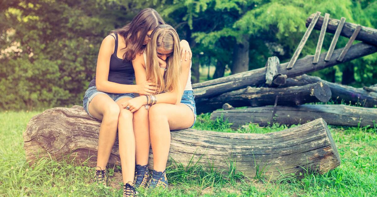 Two girls hugging as they sit on a log outside.