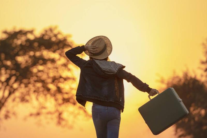 A woman walking toward the setting sun, one hand holding a hat to her head, the other swinging a small suitcase.