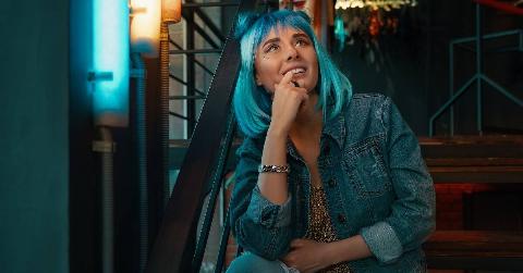 A woman with bright blue hair sitting on a staircase otuside, looking up to the sky with a hopeful expression.