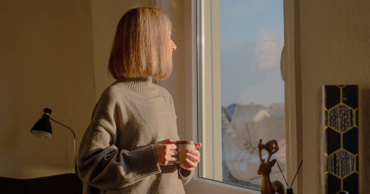 A woman holding a mug and looking out a window in her home.
