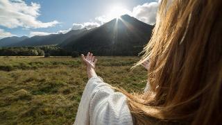 A shot taken over a woman's shoulder of her extending her arms towards the sun rising over a mountain.
