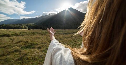 A shot taken over a woman's shoulder of her extending her arms towards the sun rising over a mountain.