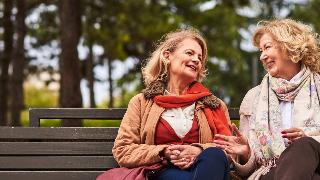 Two friends smiling as they sit and chat on a bench.