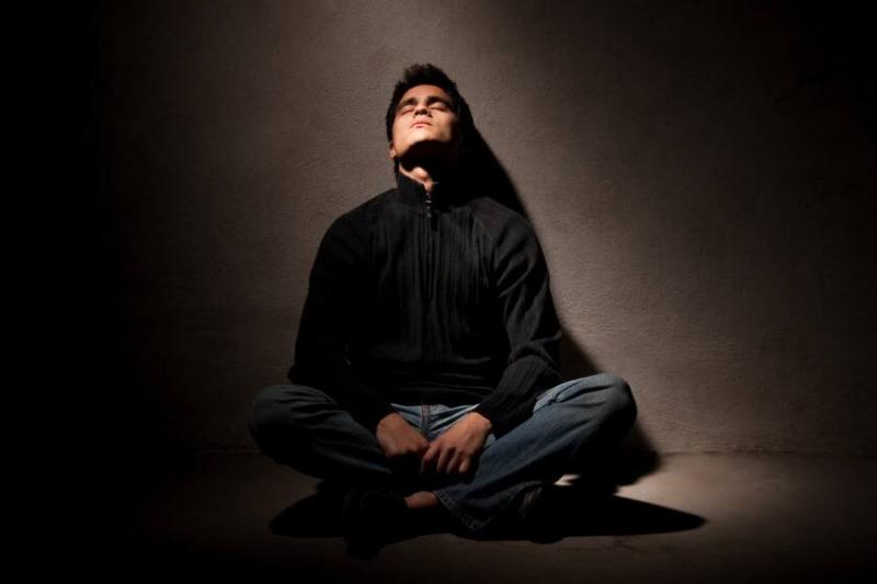 A man sitting on the ground in a spotlight, head tilted up, eyes closed.