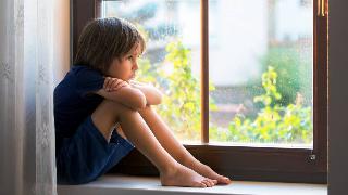 A young boy sitting in a windowsill, knees to his chest, arms on his knees, and chin on his arms, looking out the window sadly.