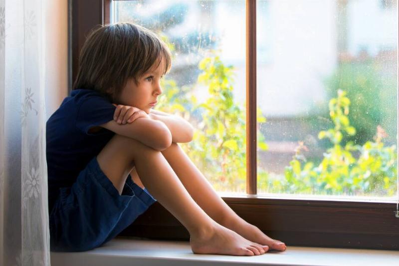 A young boy sitting in a windowsill, knees to his chest, arms on his knees, and chin on his arms, looking out the window sadly.