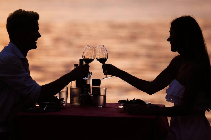 The silhouette of a couple sitting at a table in fromt of the water, clinking wine glasses together.