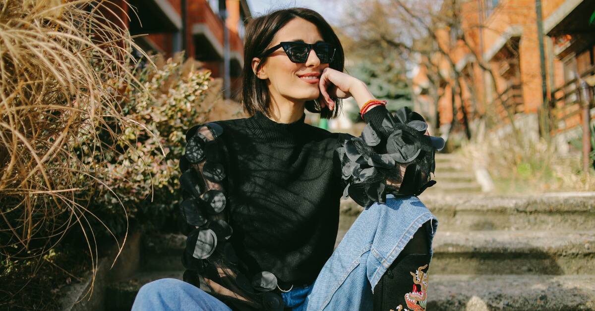 A fashionable woman in a statement shirt and funky sunglasses, smiling and posing for a photo.