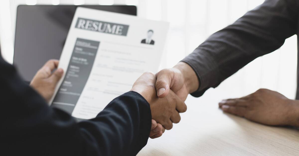 A close shot of two people shaking hands, one of them holding up a resume in the background.