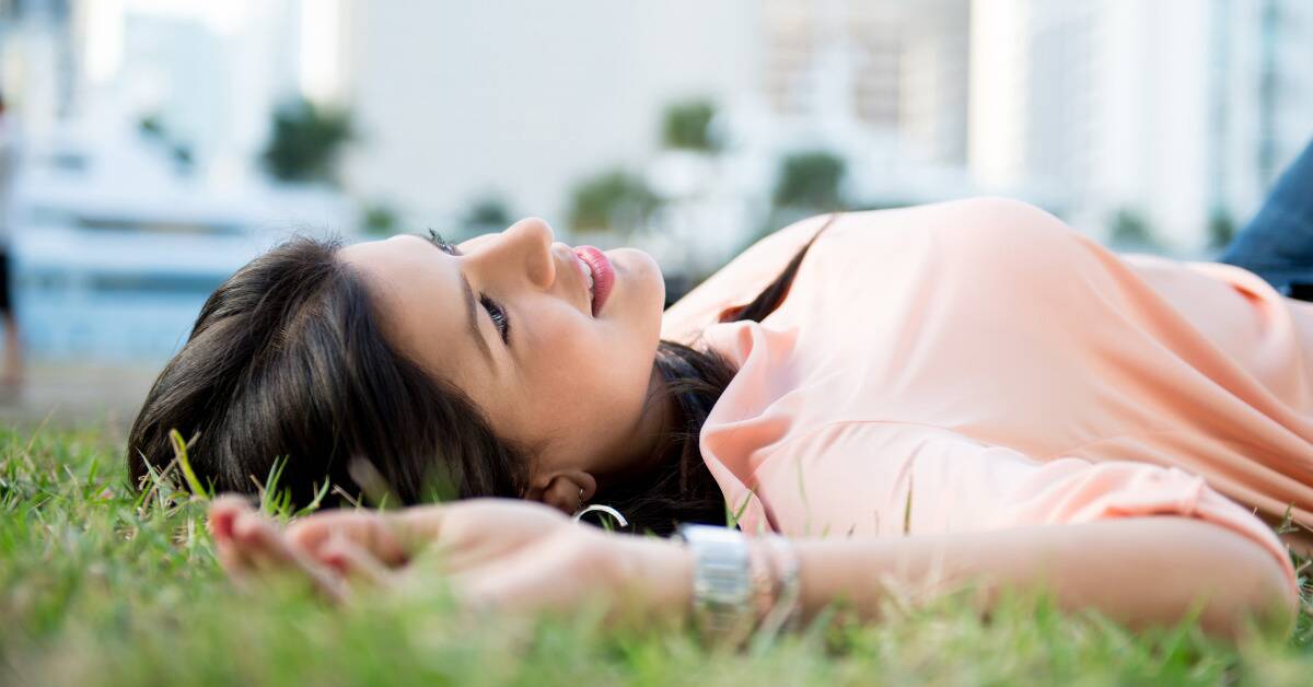 A woman laying in the grass, smiling as she watches the sky.